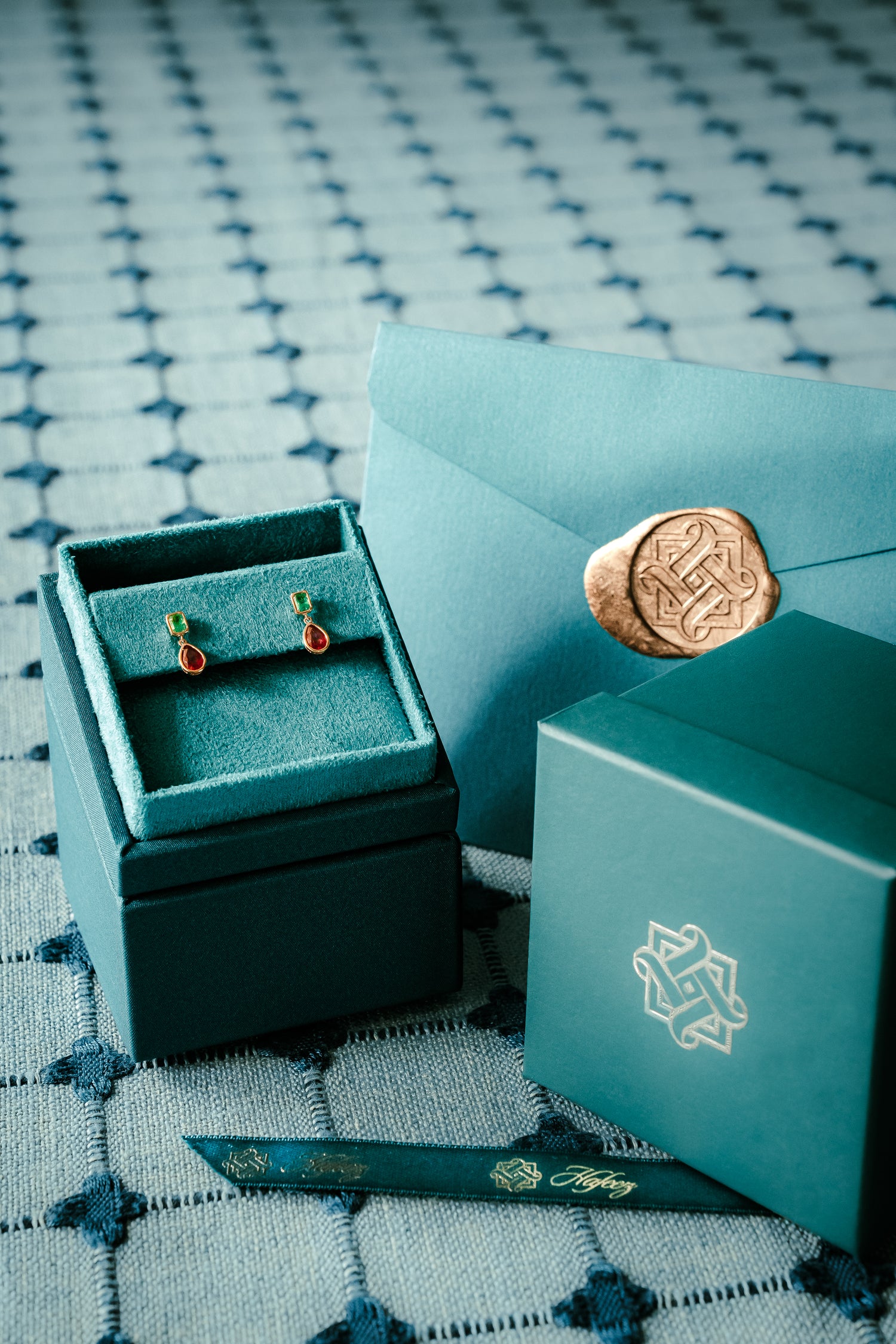 Hafeez eal jewelry box with earrings, teal envelope with wax seal, and teal box with logo on a patterned fabric background.