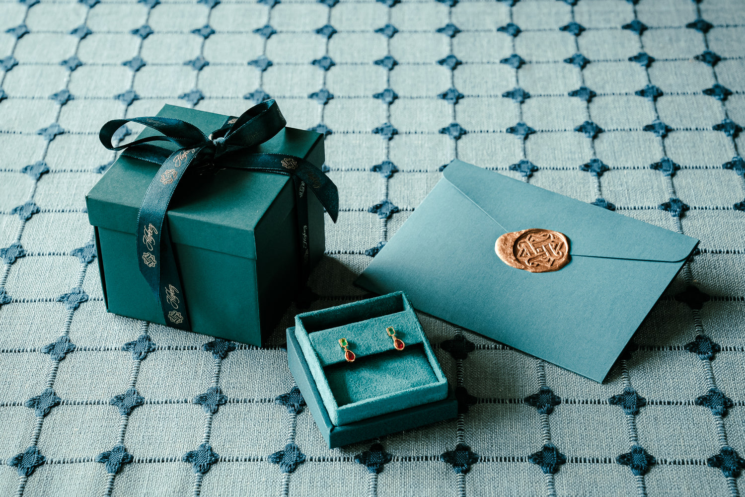 Hafeez Jewellery packaging showing a teal blue box with branded ribbon, a pair of emerald and rubies earrings in a jewellery box and an envelope with gold stamp.