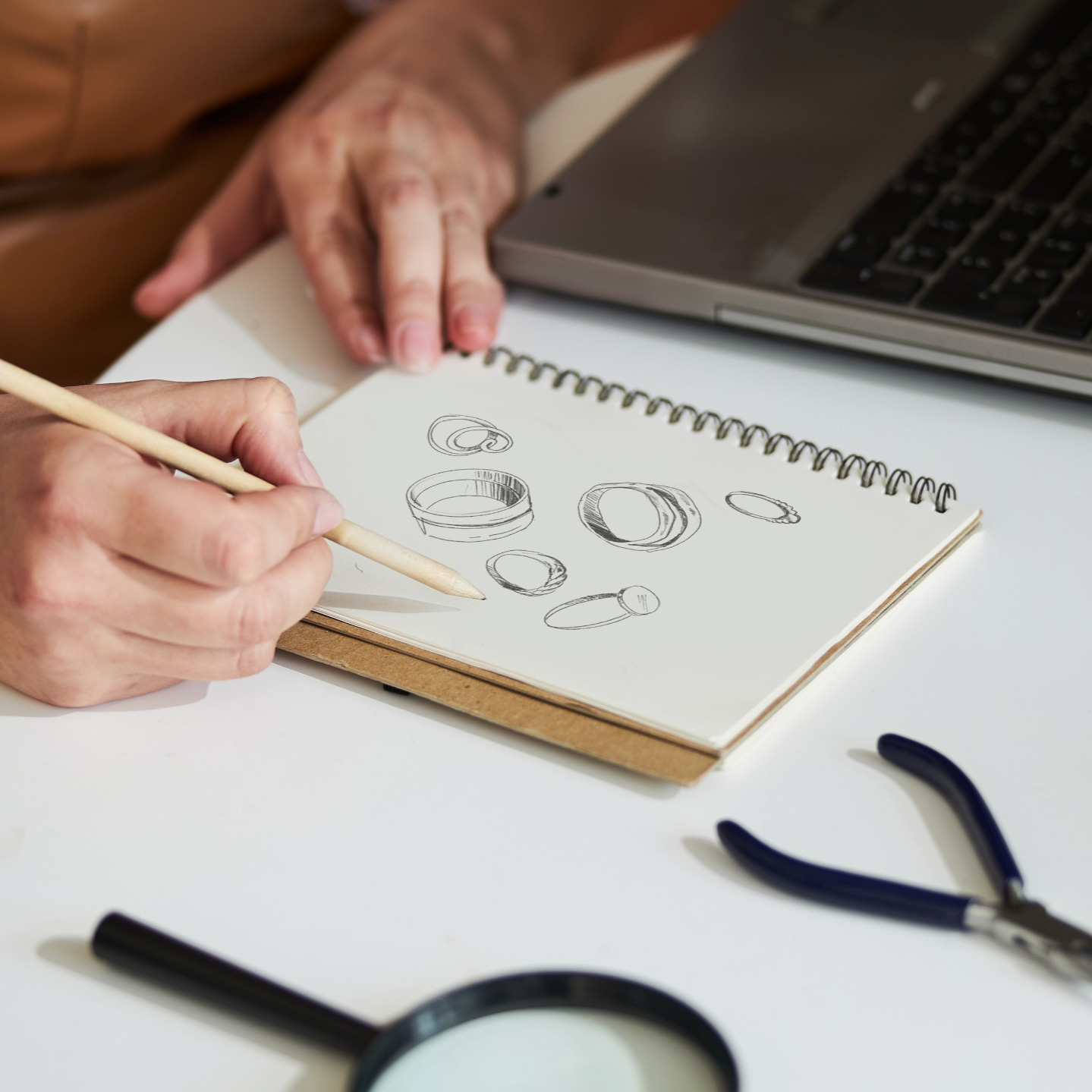 Person drawing sketches in a notebook with a laptop and magnifying glass on a table.