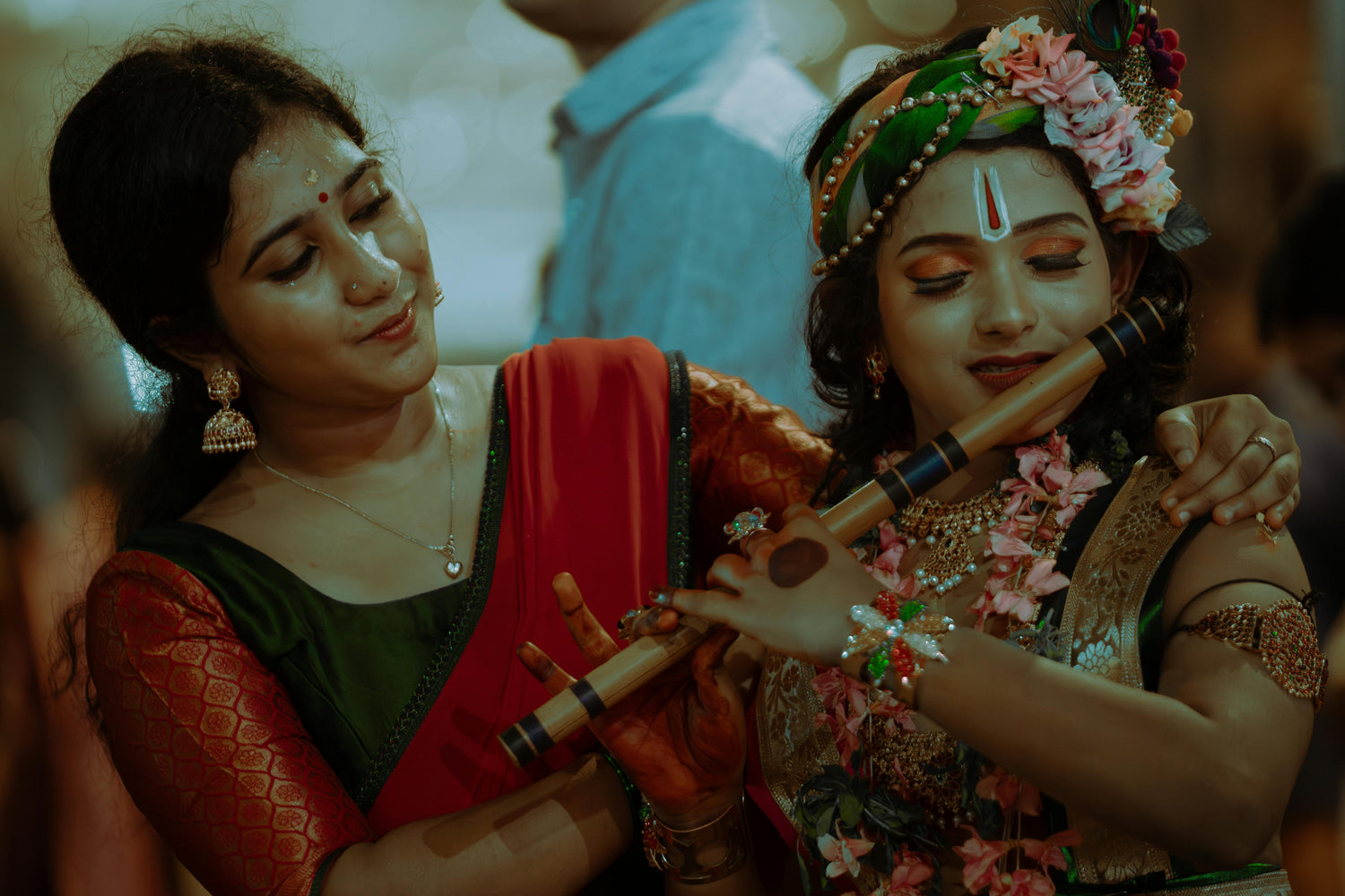 Two women, one in a red saree and the other in traditional attire with floral decorations, playing flutes.